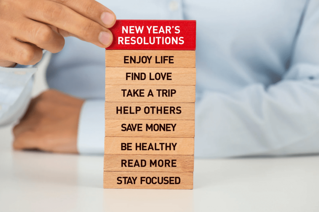 Una torre de piezas JENGA que tienen lo siguiente escrito: New year's resolutions, enjoy life, find love, take a trip, help others, save money, be healthy, read more, stay focused.
