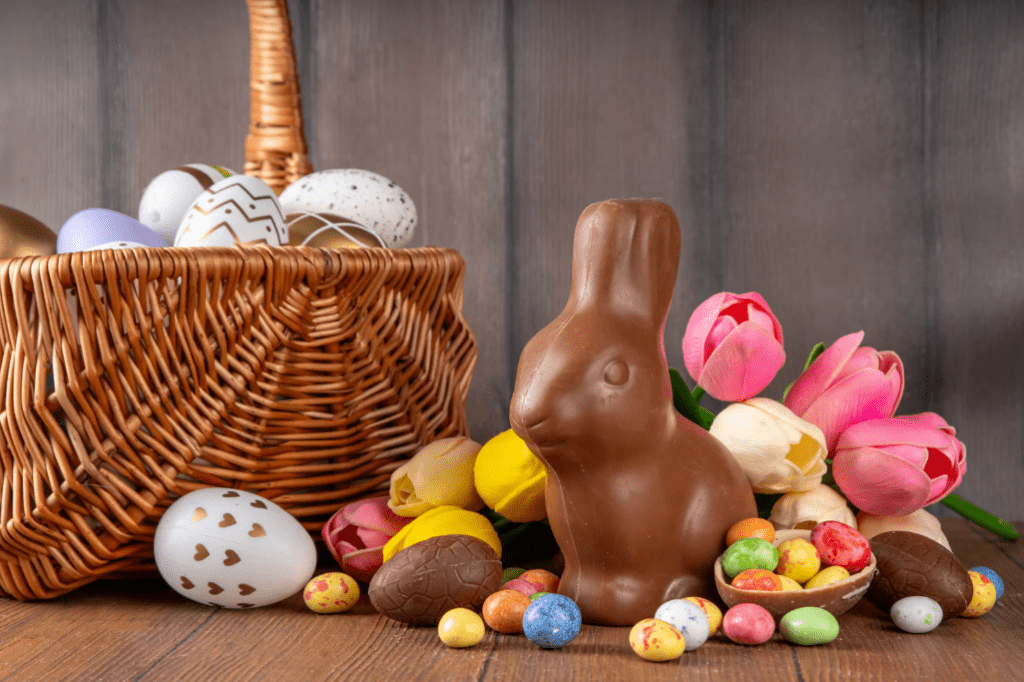 Una cesta con huevos decorados, y a su lado flores, un conejo de chocolate, y huevos de caramelo y chocolate.