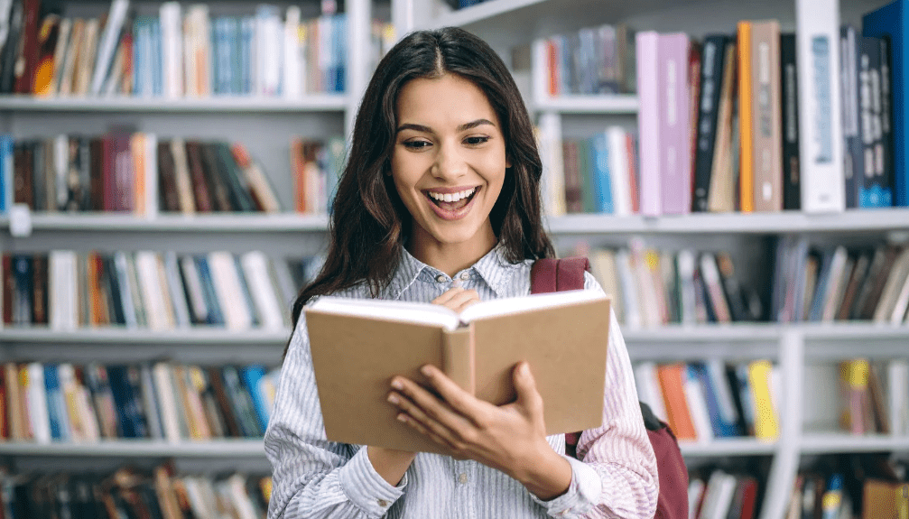 Una joven se muestra alegre frente a un libro abierto, con una estantería de libros como fondo.