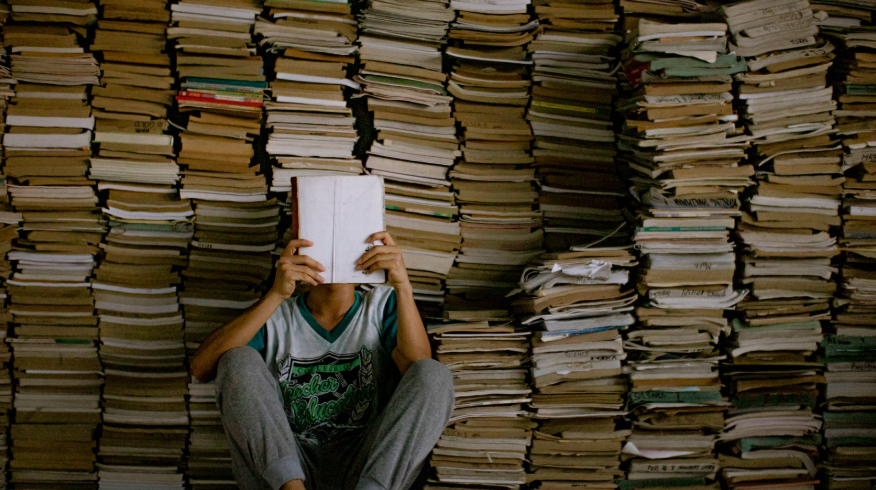 Un joven con la cara tras un libro, y pilas y pilas de libros tras él.